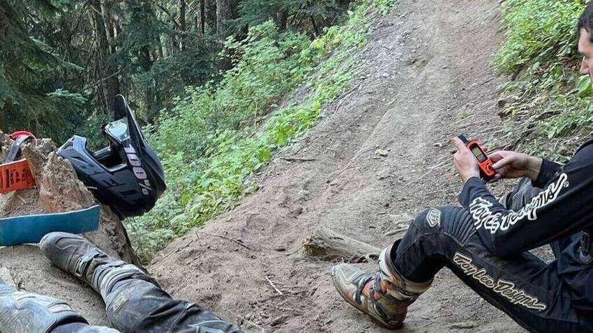 A mountain biker on a Pacific Northwest dirt trail holding an orange Garmin inReach satellite messenger, after stopping during a group ride.
