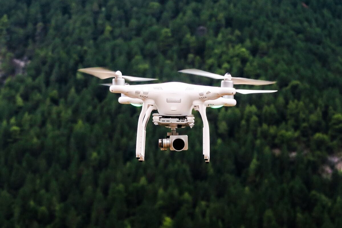 A quadcopter drone hovering above a dense evergreen forest canopy with landing lights active, illustrating the modern era of backcountry aviation.