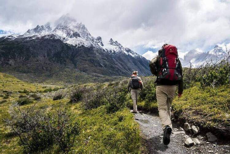 Two hikers with backpacks walking a dirt trail into a rugged mountain landscape under overcast skies.