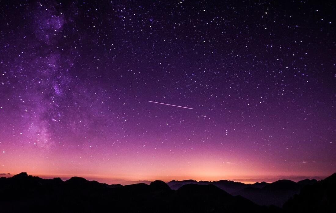 A satellite trail crossing a vivid purple Milky Way night sky above a dark mountain horizon.