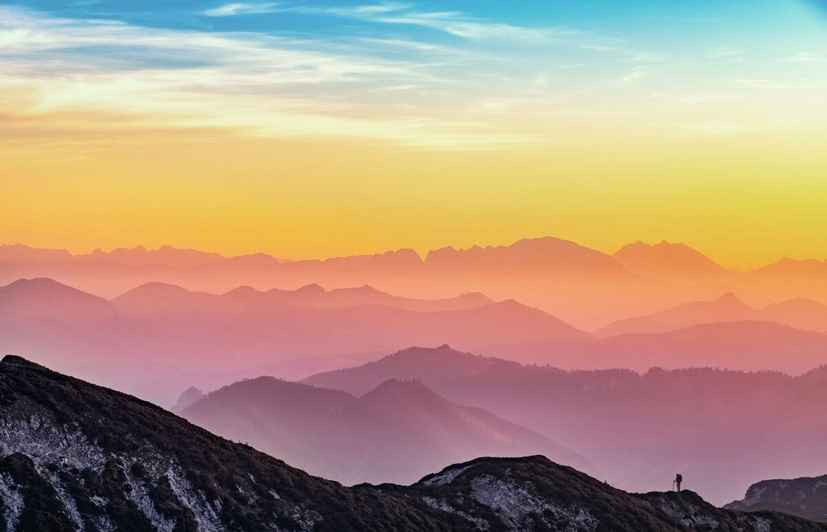 Layers of mountain ridges fading into the horizon at sunset, with a single small hiker silhouette on a rocky ridge.