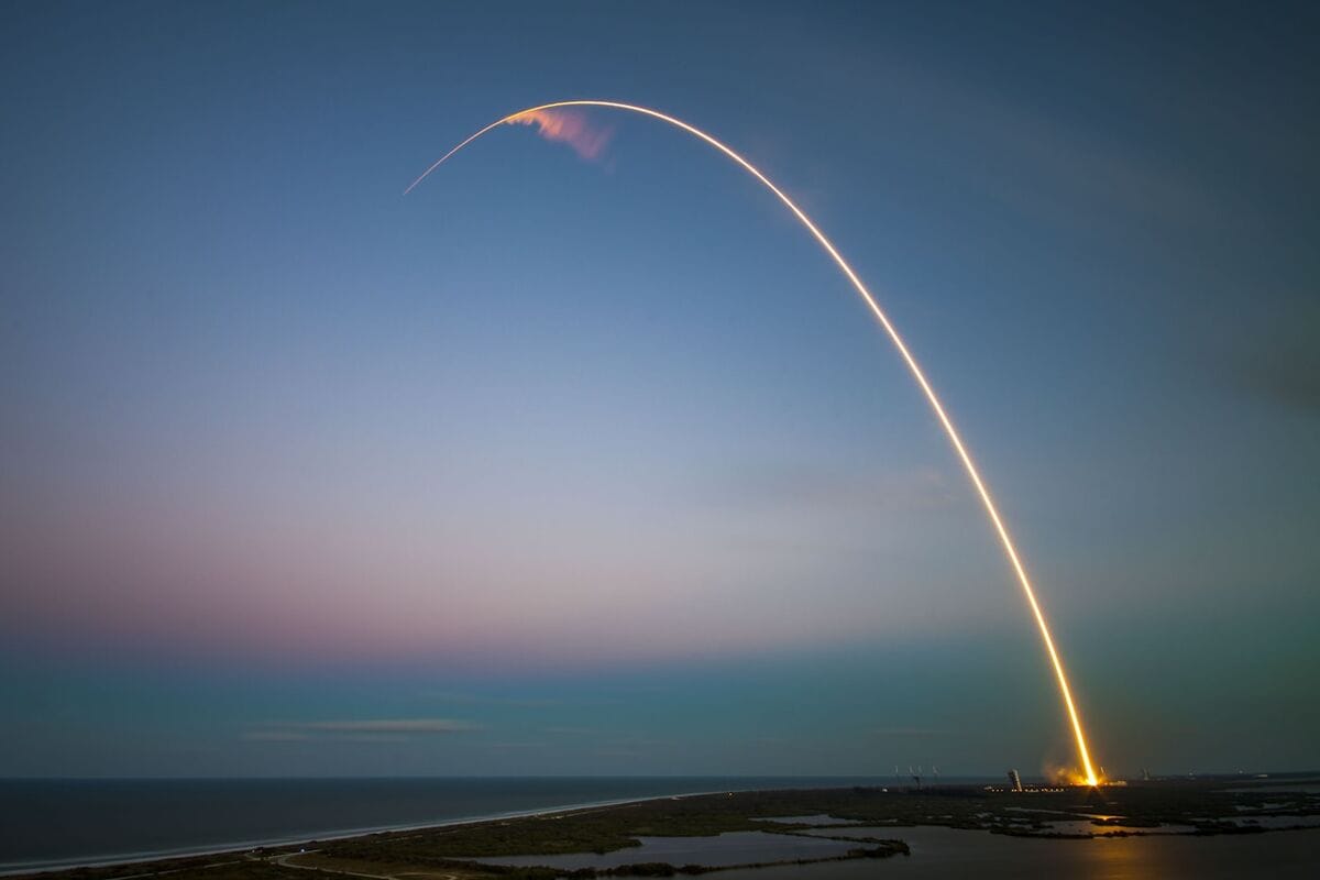 A rocket launch arc traced across a twilight sky over a coastal launch pad, the trail of light bending with the curvature of the Earth.