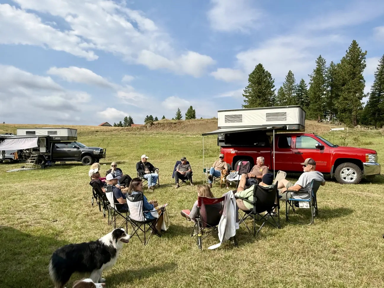 Campers sit in a circle of camp chairs in a grassy field, with a dog in the foreground and truck campers parked behind them