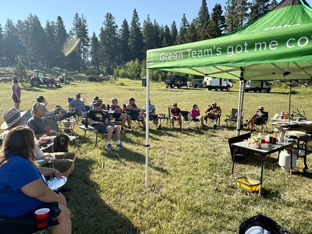 A group of campers sits in camp chairs under and around a green canopy tent in a grassy field lined with pine trees