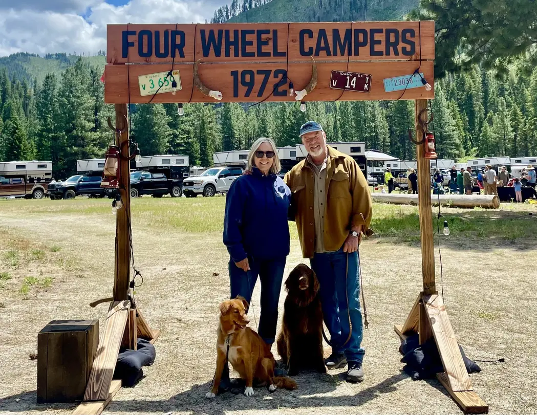 Two people and two dogs stand under a wooden Four Wheel Campers 1972 sign, with rows of pop-up truck campers parked among pine trees behind them