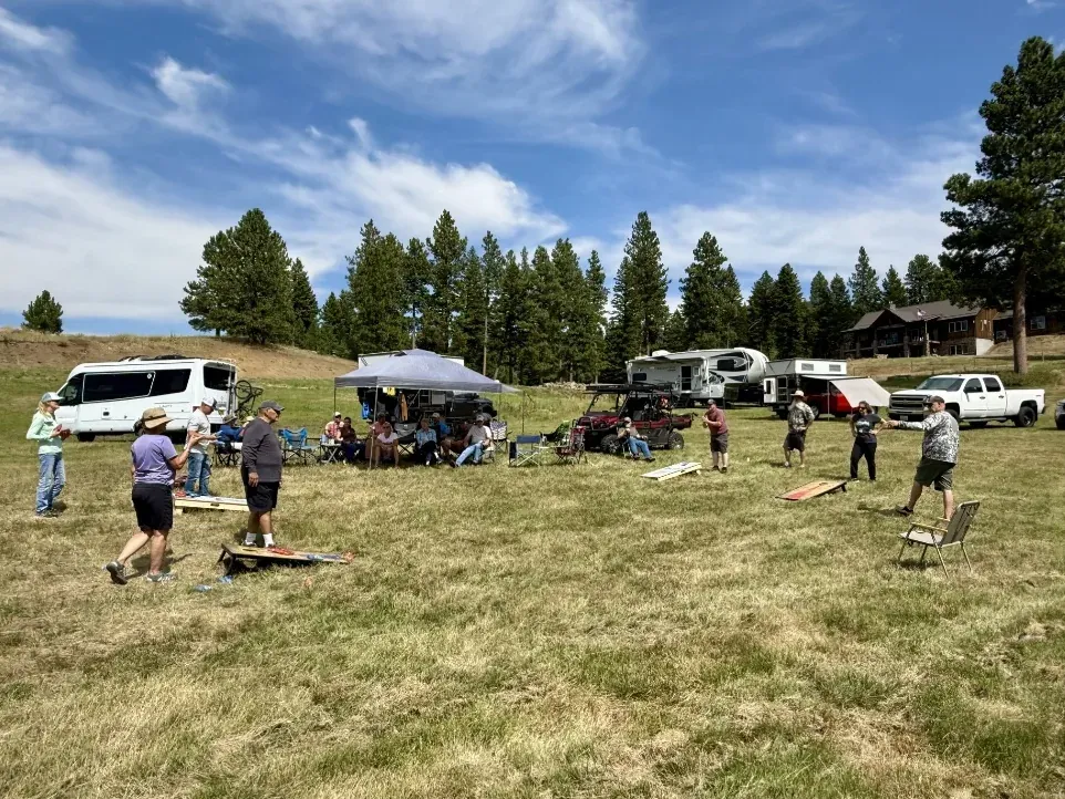 A group plays cornhole in a mowed field, with a canopy tent, vans, and truck campers parked along the pine trees behind them
