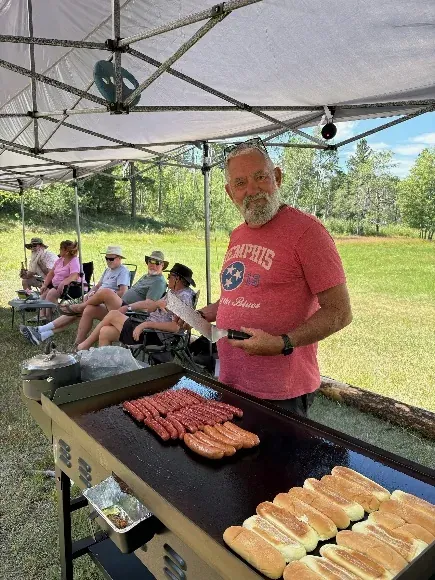 A man in a red T-shirt cooks hot dogs on a flat-top griddle under a canopy while a group sits in camp chairs behind him