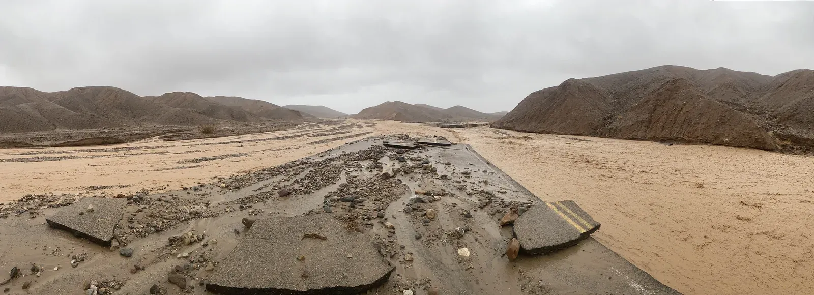 Muddy water and debris flowing over damaged Mud Canyon Road beneath storm clouds