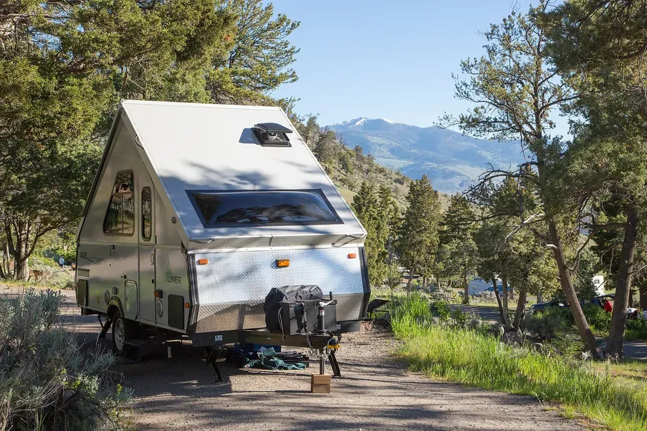 Small A-frame pop-up travel trailer parked at Mammoth Hot Springs Campground in Yellowstone with mountains behind.