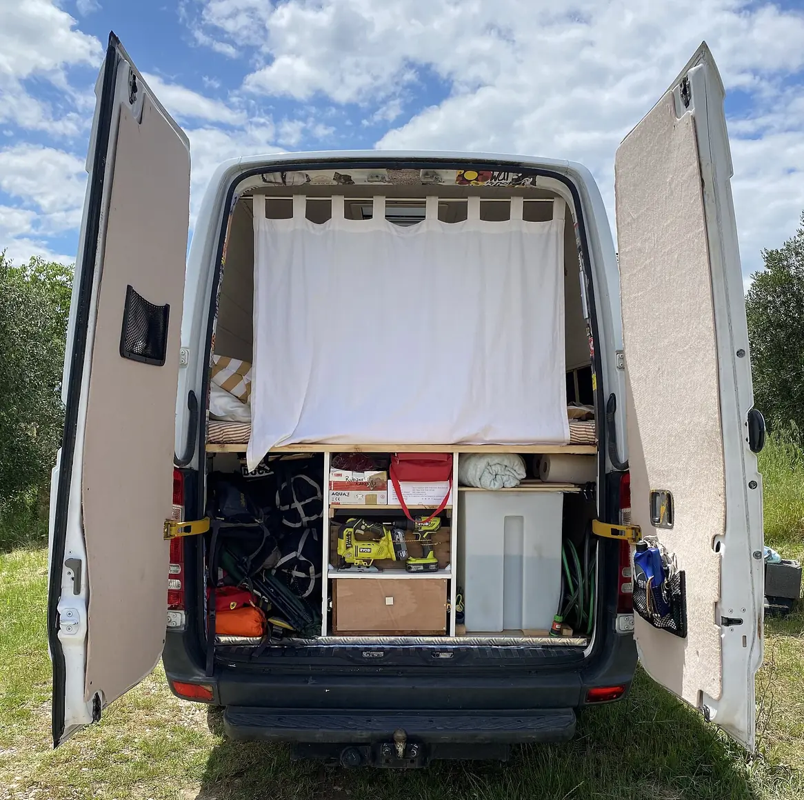 Self-converted white 2009 Mercedes Sprinter camper van with rear doors open showing a raised bed and storage underneath.