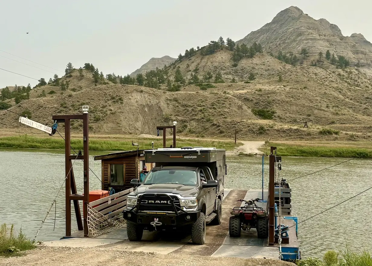 A dark pickup with a truck camper rides a cable ferry across a river, with grassy, eroded hills behind it