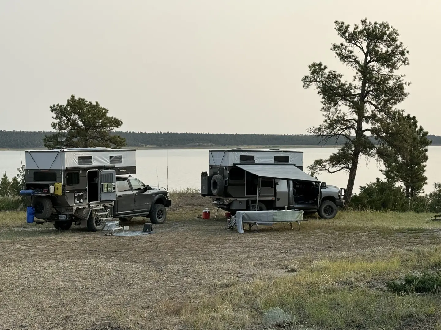 Two pop-up truck campers parked on open ground beside a lake under a hazy evening sky