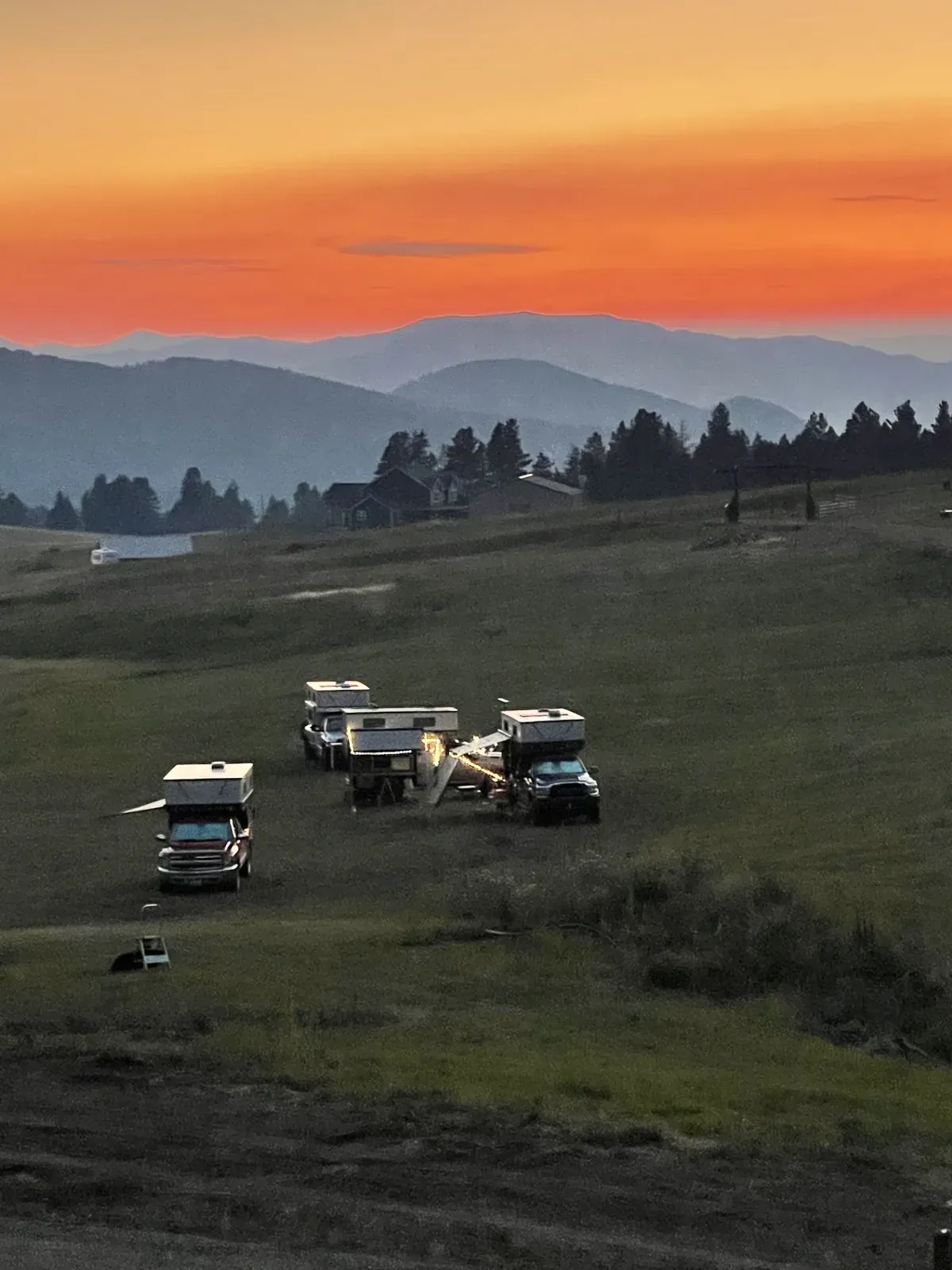 Truck campers parked on a hillside meadow at sunset, with an orange sky over distant ridgelines