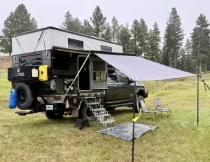 A black pickup with a pop-up truck camper and an extended side awning parked in a grassy clearing surrounded by pine trees